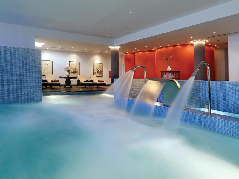 Basen kryty z niebieskimi płytkami, fontannami wodnymi i przytulną częścią wypoczynkową w tle.