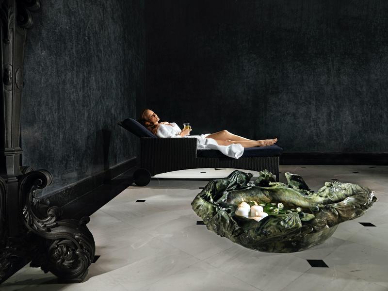 Osoba odpoczywa na leżaku w ciemnym pokoju z dekoracyjnym zielonym obiektem na podłodze.