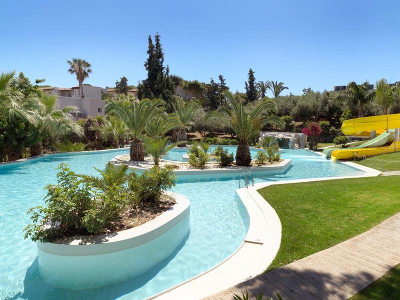 Basen rekreacyjny z wysepkami wodnymi, otoczony palmami i żółtą zjeżdżalnią wodną.