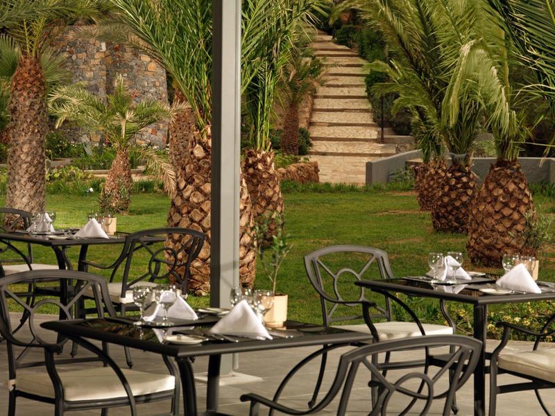 Zewnętrzny teren hotelu z stołami, krzesłami, palmami i schodami w tle.