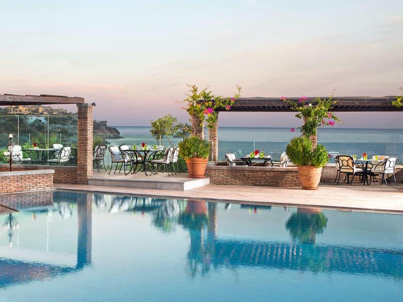 Hotelowy basen z widokiem na morze i meblami ogrodowymi o zachodzie słońca.