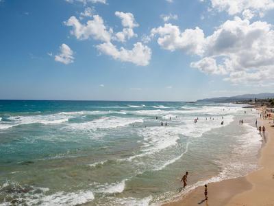 Spiaggia sabbiosa con persone che nuotano e camminano lungo la riva in una giornata di sole.
