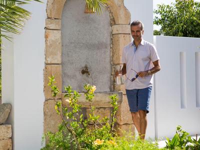 Uomo con camicia bianca e pantaloncini blu vicino a muro di pietra e piante davanti a casa bianca.