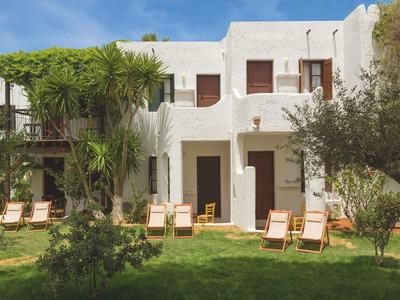 Weißes Gebäude mit braunen Türen und Fenstern, umgeben von grünen Bäumen und Liegestühlen.