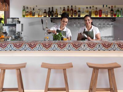 Dos camareros preparando bebidas detrás de una barra decorativa con tres taburetes de madera.