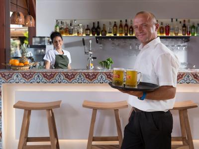 Cameriere sorridente che porta bevande su un vassoio in un bar moderno con sgabelli e un barista sullo sfondo.