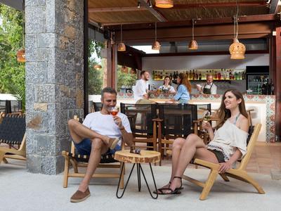 Pareja relajándose en sillas plegables frente a un bar abierto con barman al fondo.