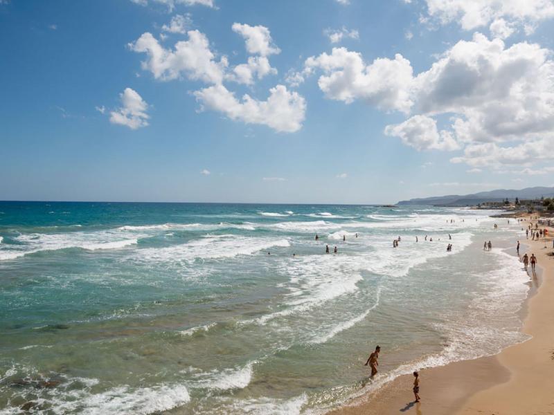 Spiaggia sabbiosa con persone che nuotano e camminano lungo la riva in una giornata di sole.