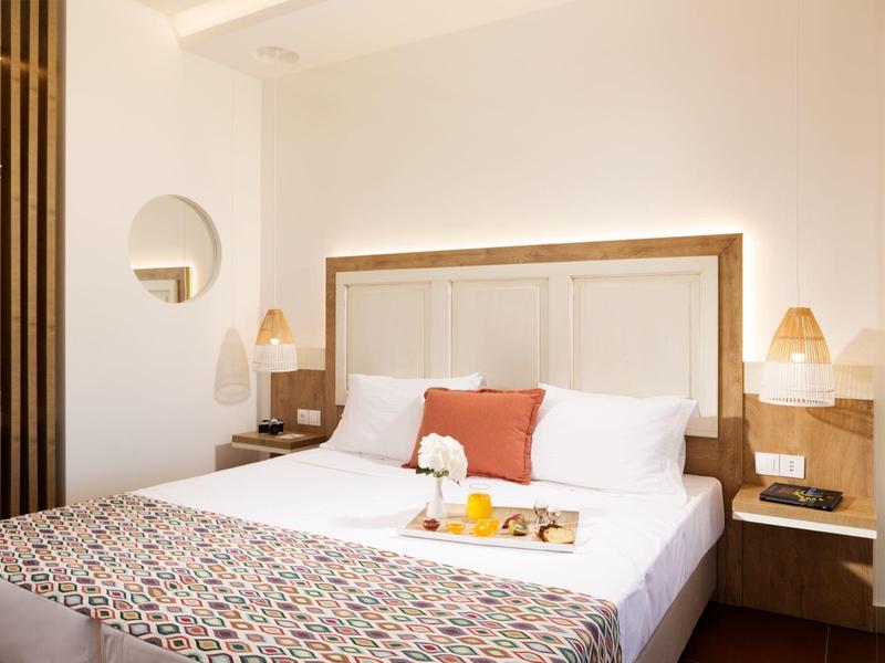 Camera d'hotel luminosa con grande letto, cuscini bianchi e arancioni, e dettagli in legno.