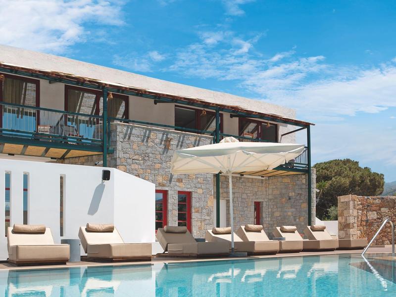 Piscina soleggiata in resort con lettini e edificio moderno in pietra sotto cielo blu.