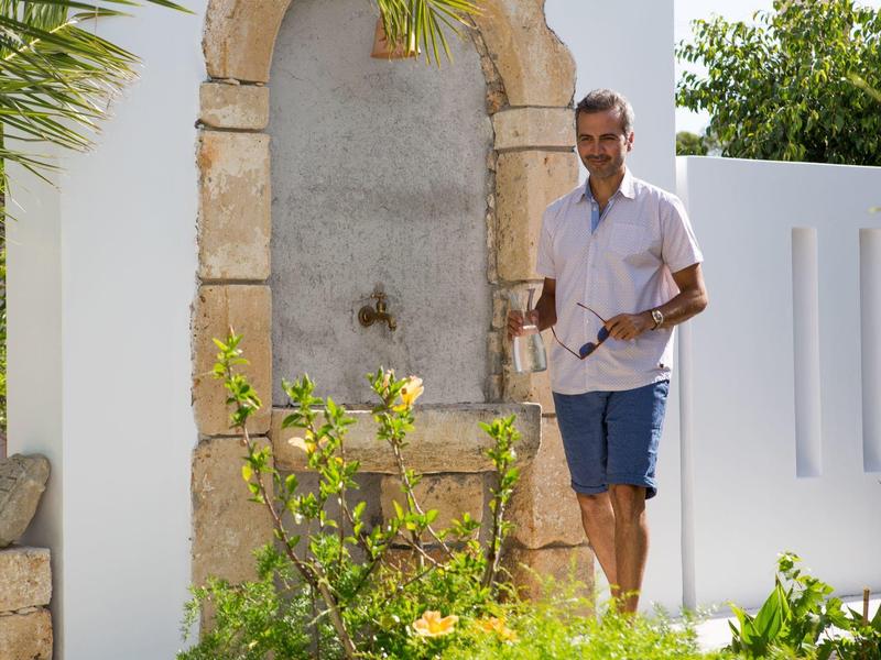 Uomo con camicia bianca e pantaloncini blu vicino a muro di pietra e piante davanti a casa bianca.
