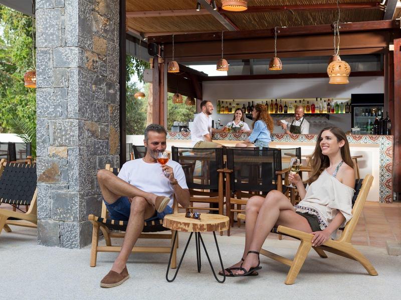 Coppia si rilassa su sedie di legno con bevande in un bar all'aperto con pilastri in pietra e vegetazione.