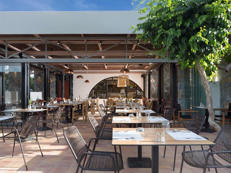 Area ristorante all'aperto con tavoli e sedie sotto un tetto parzialmente coperto e vegetazione.