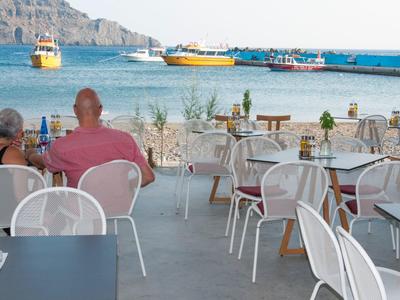 Vista su tavoli esterni di un caffè vuoti e occupati con vista sul mare e barche al molo.