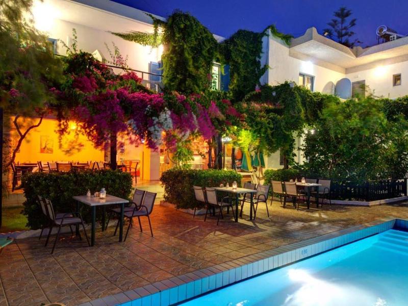 Terrazza serale di un hotel con tavoli, sedie, piante fiorite e piscina illuminata.