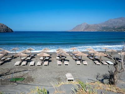 Leerer Strand mit Reihen von Sonnenliegen und Klappstühlen vor ruhigem Meer und Bergen.