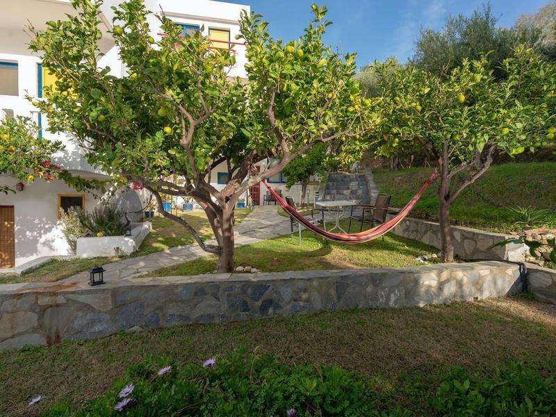 Garten mit grünen Bäumen, roter Hängematte, Steinmauer und weißem Haus im Hintergrund bei blauem Himmel.