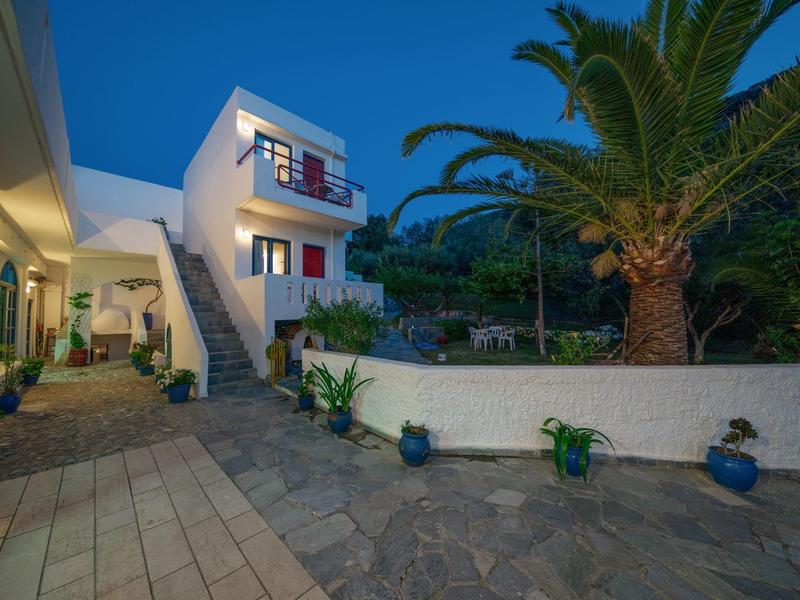 Moderne weiße Villa mit Terrasse, Treppe und Palmen im Abendlicht. Pflanzgefäße und Beleuchtung sichtbar.
