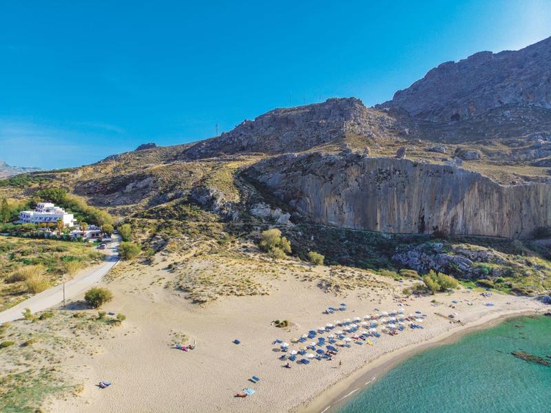 Strand mit klarem Wasser, Sonnenschirmen und bergiger, felsiger Landschaft im Hintergrund.