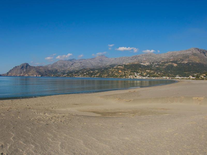 Breiter Strand mit Blick auf ruhiges Meer und Berge im Hintergrund unter blauem Himmel