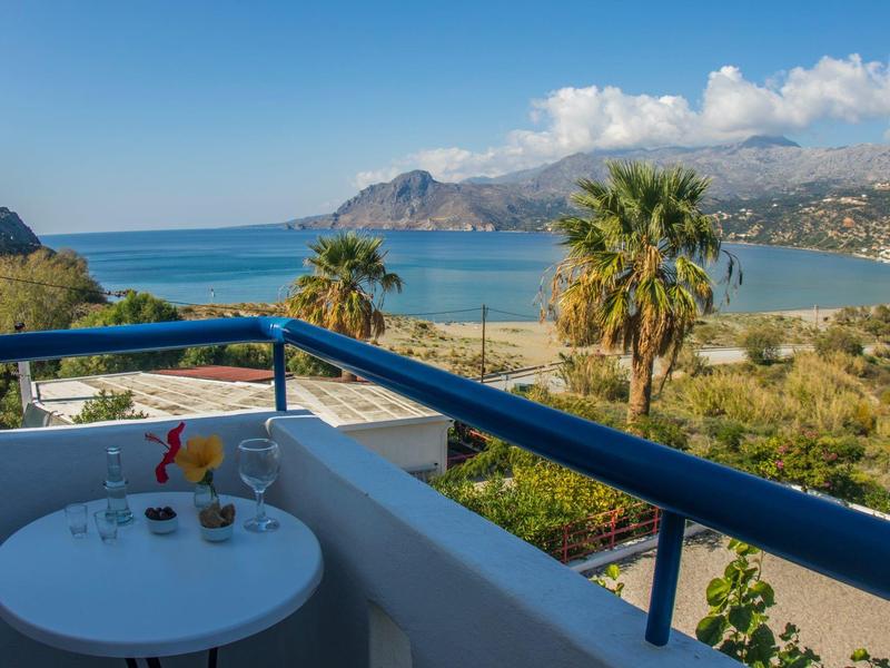 Balkon mit kleinem Tisch, Blick auf Palmen, Berge und Meer unter blauem Himmel.