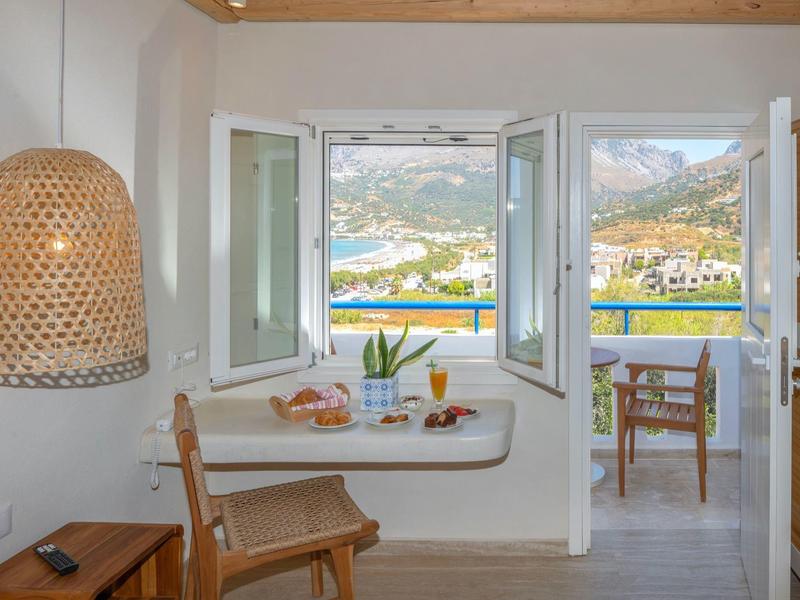 Helles Zimmer mit offenem Fenster, Blick auf Berge, Meer und Terrasse mit Stühlen.