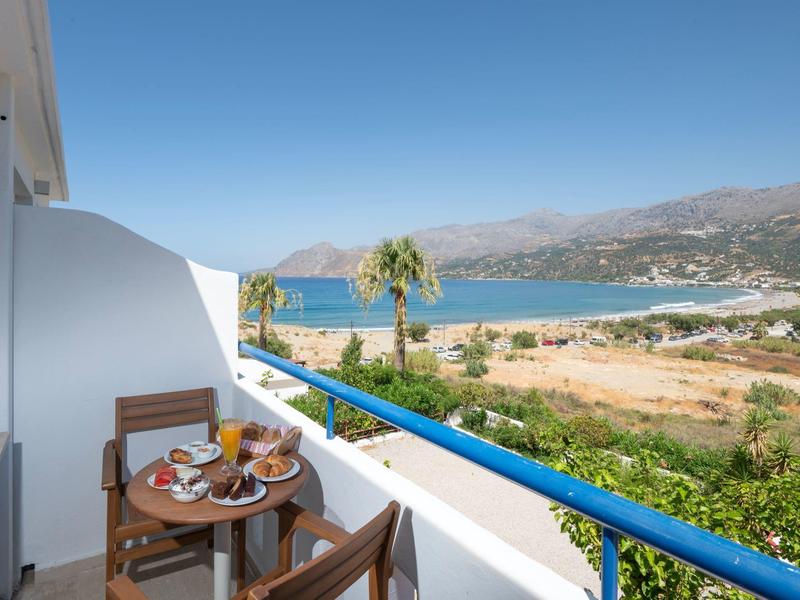 Balkon mit Holztisch und Stühlen, Frühstück und Meerblick unter klarem Himmel.