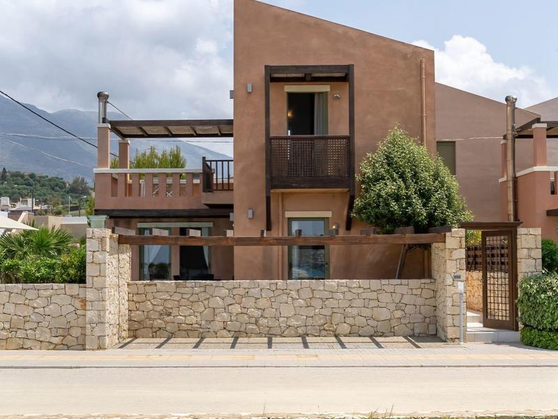 Casa vacanze moderna con terrazza e muro di pietra davanti a prato verde sotto cielo azzurro.