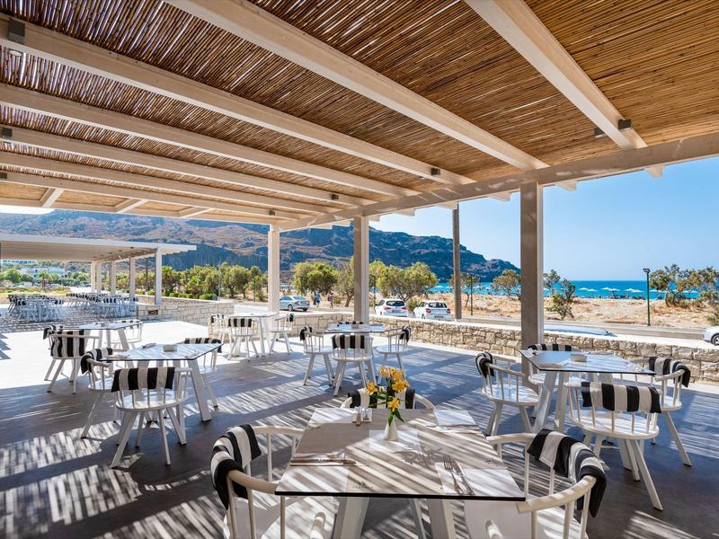 Area coperta aperta con tavoli e sedie, vista su spiaggia e montagne.