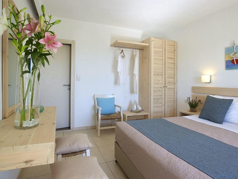 Chambre d'hôtel lumineuse avec lit, armoire, chaise et vase avec des fleurs sur une table en bois.
