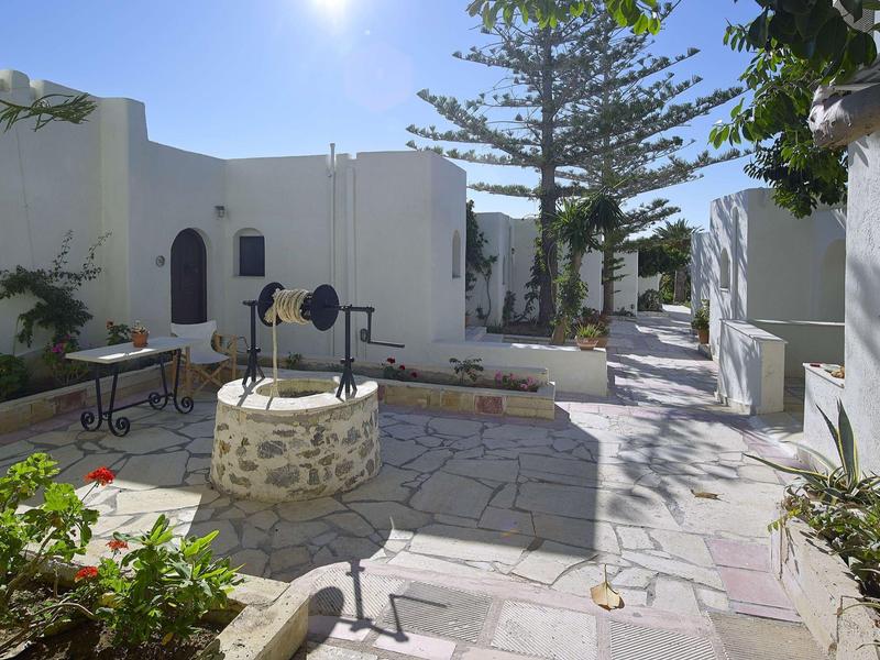 Cour avec des bâtiments peints en blanc, un puits et un sol en pierre sous un soleil éclatant.