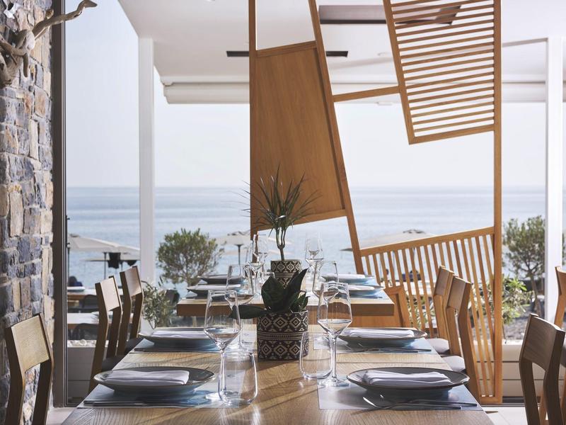 Coin repas avec table en bois, chaises et vue sur la mer à travers de grandes fenêtres.