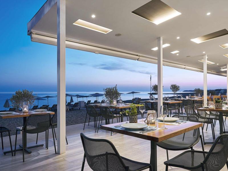 Restaurant en plein air avec vue sur la mer au crépuscule, tables dressées et éclairage moderne.