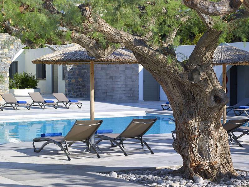 Espace piscine avec chaises longues sous de grands arbres et murs en pierre en arrière-plan.