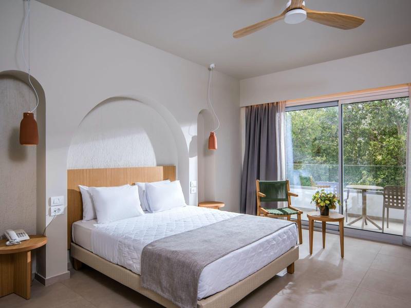 Hell eingerichtetes Schlafzimmer mit Doppelbett, Deckventilator, Sitzbereich und großen Fensterfront.