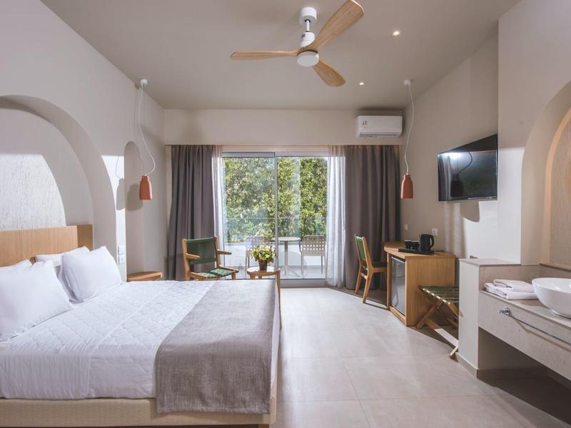 Helles Hotelzimmer mit Doppelbett, Deckenventilator, Balkon und moderner Einrichtung in neutralen Farben.