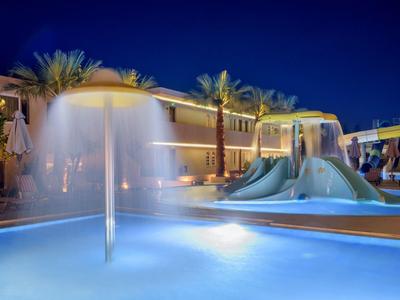 Oświetlony nocą basen hotelowy z atrakcjami wodnymi i zjeżdżalniami, otoczony palmami i miejscami do siedzenia.