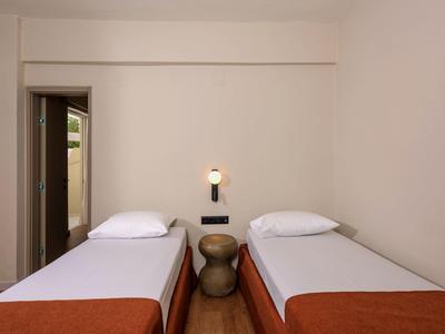 Prosto urządzone pokoje hotelowe z dwoma pojedynczymi łóżkami, stolikiem nocnym i lampą ścienną.