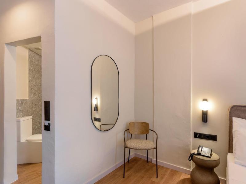 Nowoczesny pokój hotelowy z jasną drewnianą podłogą, krzesłem, owalnym lustrem i lampką nocną.