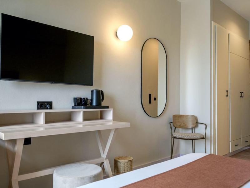 Nowoczesny pokój hotelowy z telewizorem na ścianie, lustrem, krzesłem i biurkiem w neutralnych kolorach.