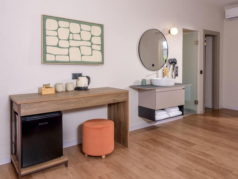 Nowoczesna część pokoju hotelowego z biurkiem, mini lodówką, stołkiem i umywalką na ścianie.