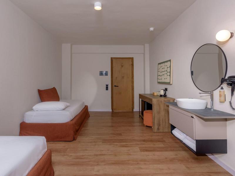 Prosty pokój hotelowy z dwoma łóżkami, drewnianą podłogą i nowoczesną umywalką na ścianie.