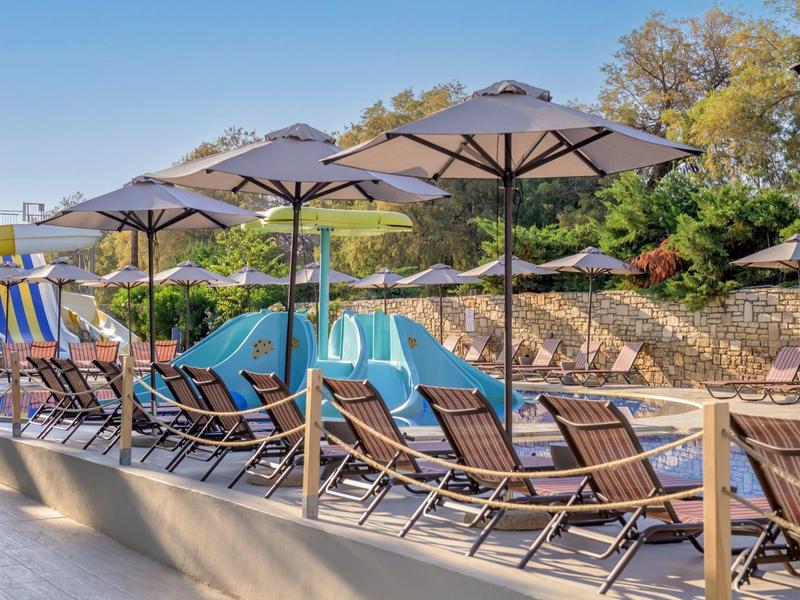 Drewniane leżaki z niebieskimi poduszkami pod parasolami przy basenie w słonecznym otoczeniu.