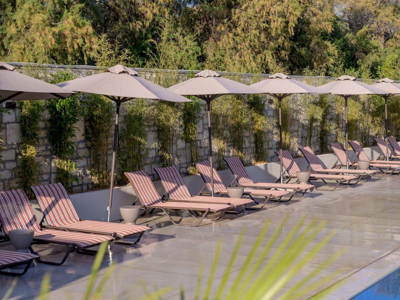 Rząd leżaków z parasolami przy basenie otoczonym zielonymi drzewami.