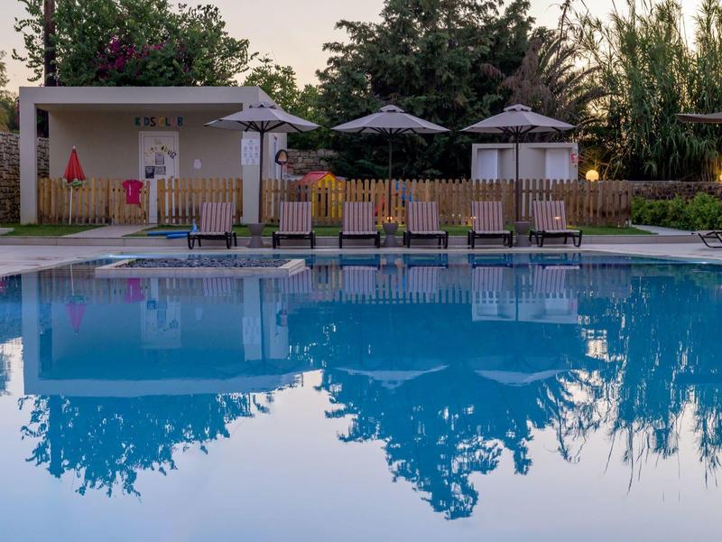 Pusty basen z leżakami, parasolami i małym budynkiem w tle.