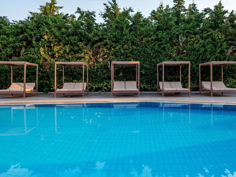 Pięć wygodnych leżaków z daszkiem przed zielonym żywopłotem przy niebieskim basenie.