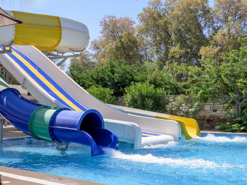 Kilka zjeżdżalni wodnych kończy się w basenie otoczonym zielonymi drzewami w słoneczny dzień.