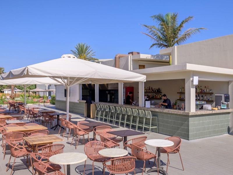 Teren zewnętrzny hotelu z kawiarnią, stołami, krzesłami i parasolami pod czystym niebem.