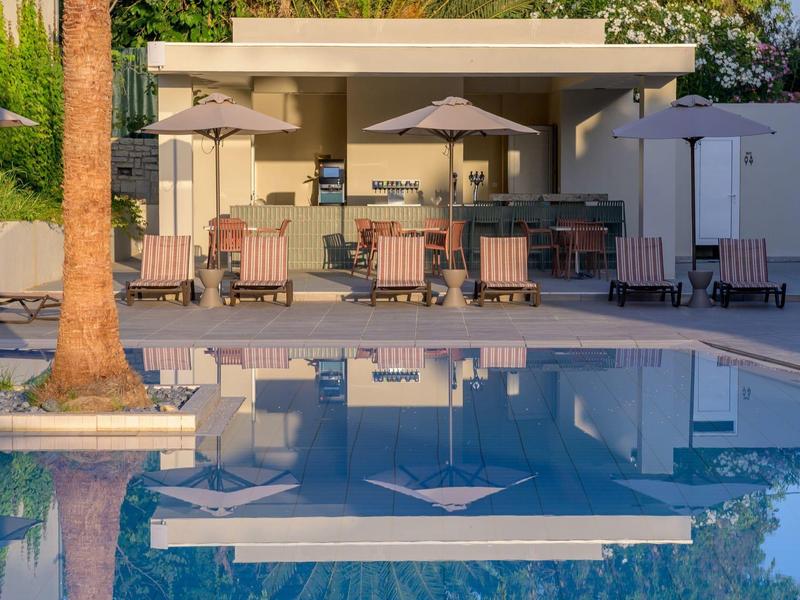 Spokojny basen z krzesłami, stołami i parasolami przed budynkiem o zachodzie słońca.