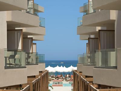 Uitzicht tussen hotelgebouwen op een zwembad met parasols en de zee op de achtergrond.